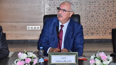 Man in a blue suit and red tie sits at a conference table with a microphone; Arabic nameplate and pink-white flowers on the table.