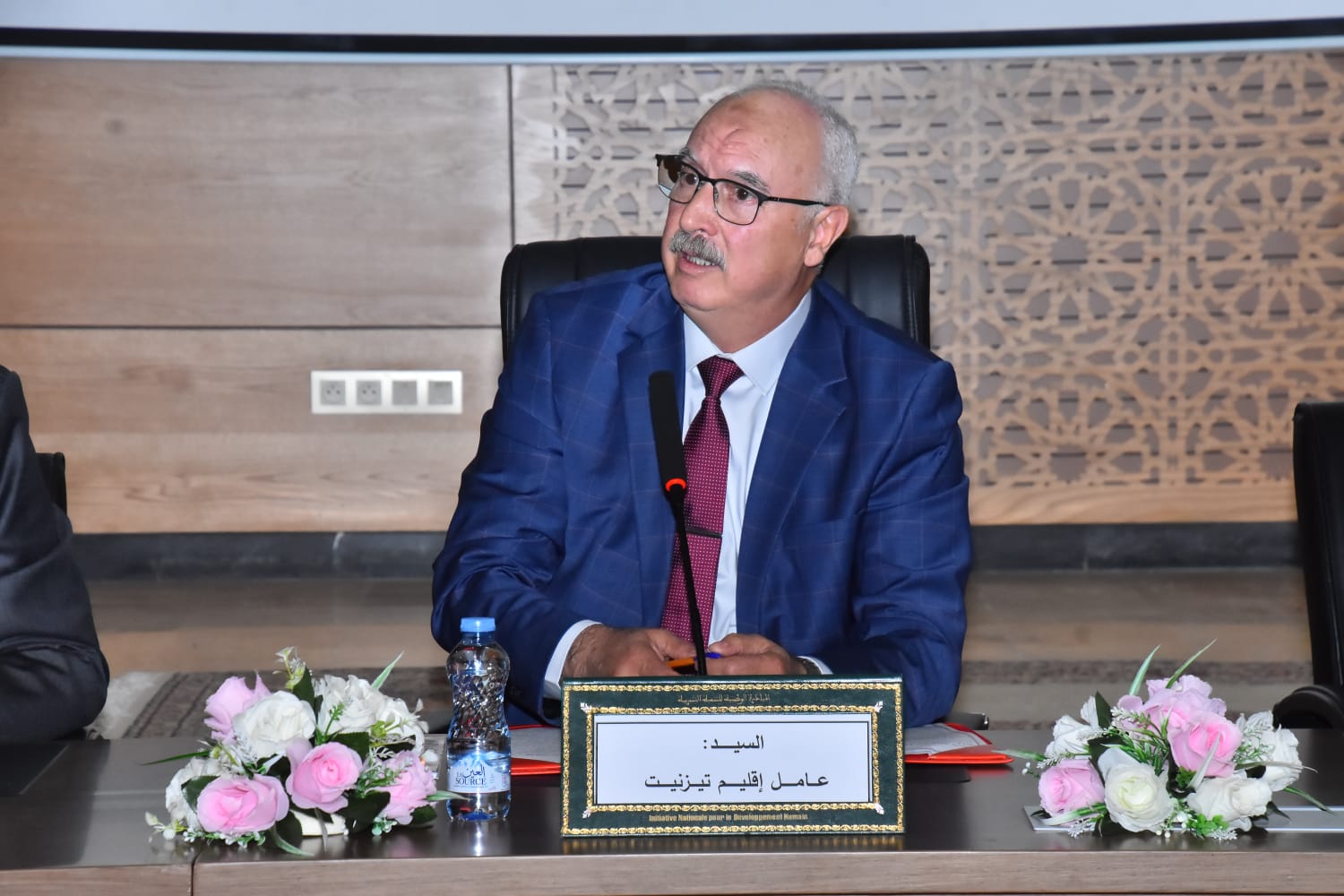 Man in a blue suit and red tie sits at a conference table with a microphone; Arabic nameplate and pink-white flowers on the table.
