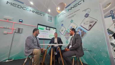 Three men in business attire sit around a table at a MEDCONNECT booth, discussing healthcare tech: a trade-show setting.