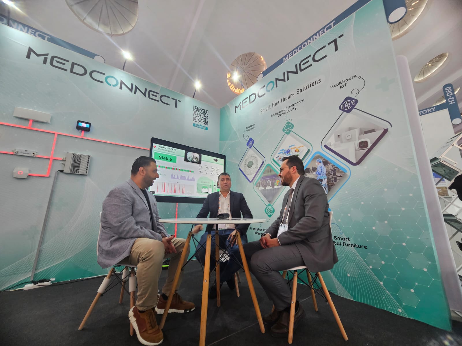 Three men in business attire sit around a table at a MEDCONNECT booth, discussing healthcare tech: a trade-show setting.