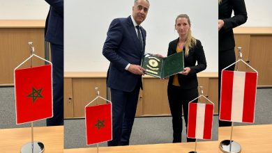 Business professionals pose with an open green folder during a formal ceremony; Moroccan and Austrian flags flank them.