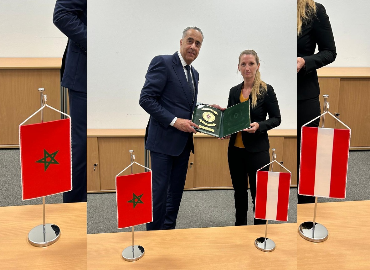 Business professionals pose with an open green folder during a formal ceremony; Moroccan and Austrian flags flank them.