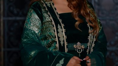 Elegant woman in a dark emerald green, embroidered gown with silver trim and statement earrings, standing with hands clasped.