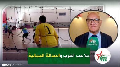WhatsApp Image 2026-05-12 at 100910 | MCG24 | موقع إلكتروني مغربي شامل متجدد على مدار الساعة News thumbnail showing an indoor futsal match with a yellow-jersey goalkeeper (#1) and players, plus a circular inset of a man in a suit giving an interview with a green microphone. Arabic headline bar at bottom.
