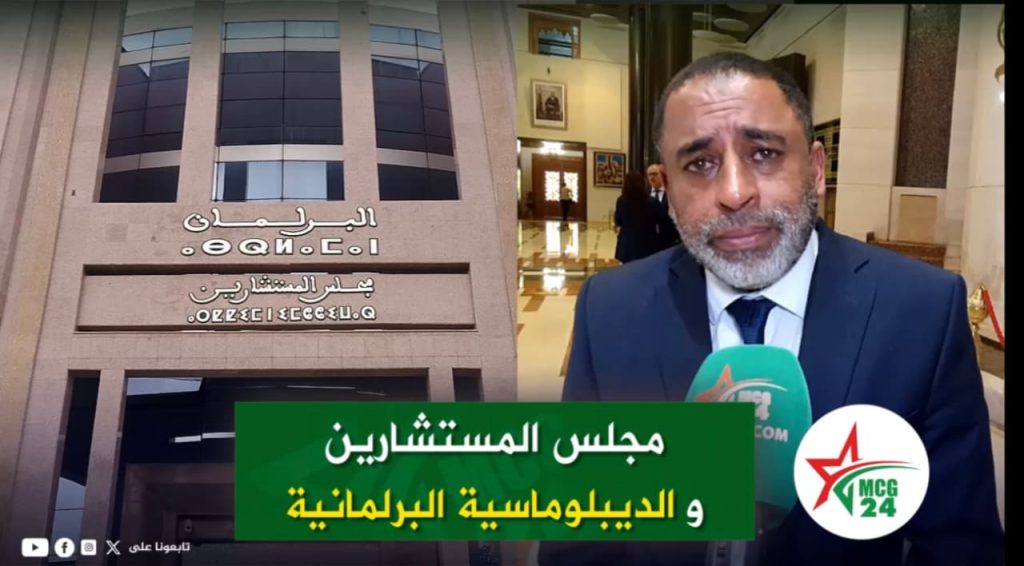 عبد القادر الكيحل: الدبلوماسية البرلمانية تعزز الدفاع عن قضية الصحراء داخل المنتديات الدولية News interview: man in a navy suit speaks into a green MC G24 microphone in a building lobby with Arabic signage on the wall behind him (about the Council of Advisors and parliamentary diplomacy).