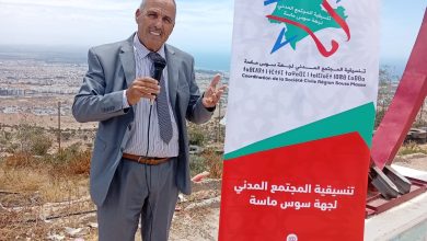 Man in a gray suit holding a microphone stands beside a tall red-and-white banner with Arabic text, outdoors overlooking a city and sea neutered by a blue sky.