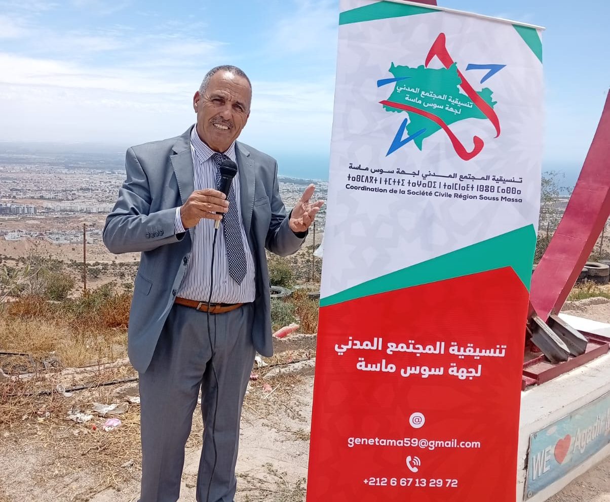 WhatsApp Image 2026-05-13 at 143541 | MCG24 | موقع إلكتروني مغربي شامل متجدد على مدار الساعة Man in a gray suit holding a microphone stands beside a tall red-and-white banner with Arabic text, outdoors overlooking a city and sea neutered by a blue sky.