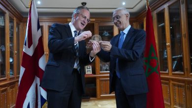 Two men in suits exchange a circular coin award, smiling, in a formal room with flags and wooden cabinets behind them