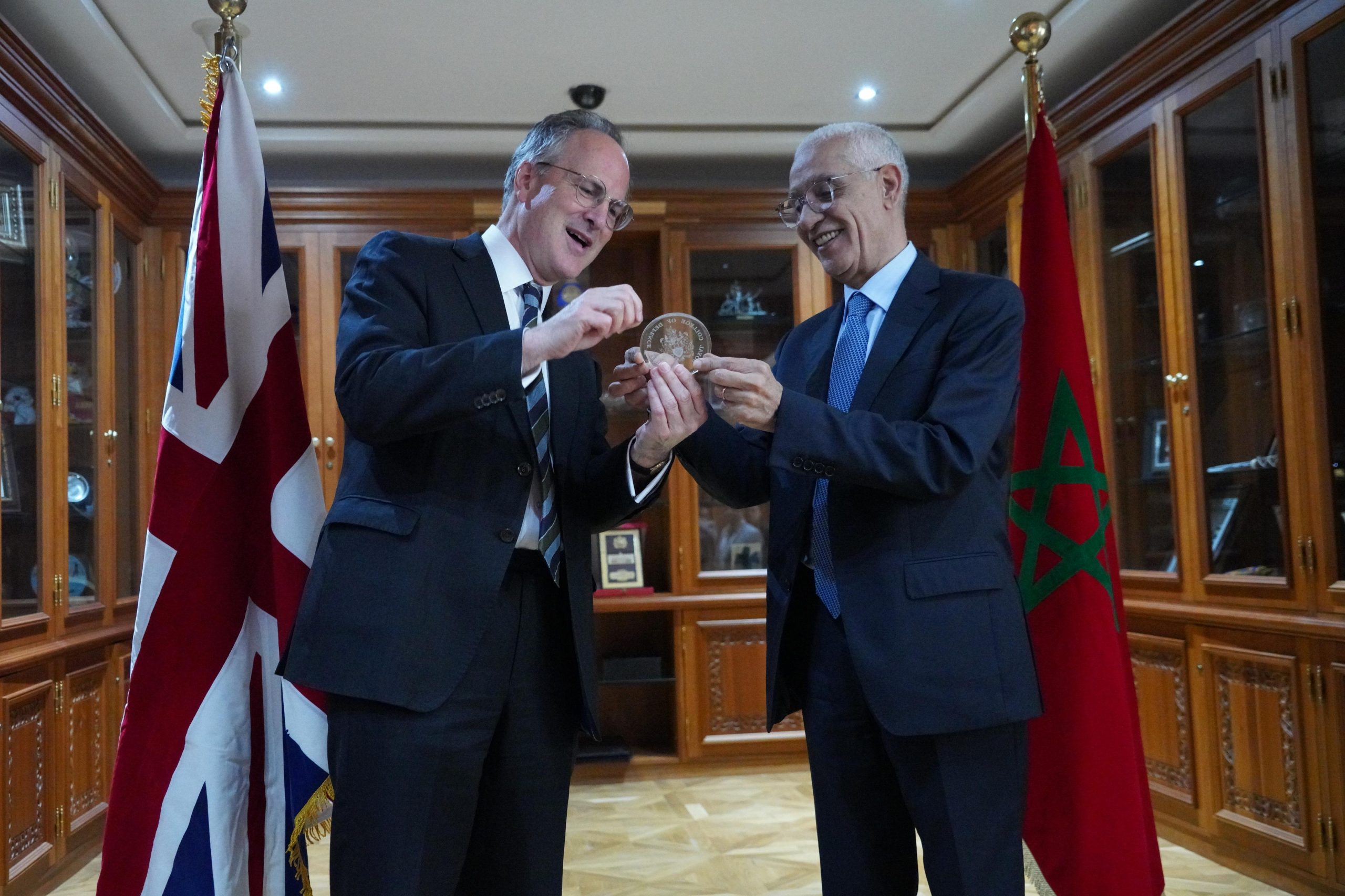 Two men in suits exchange a circular coin award, smiling, in a formal room with flags and wooden cabinets behind them