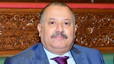 WhatsApp Image 2026-05-14 at 090203 | MCG24 | موقع إلكتروني مغربي شامل متجدد على مدار الساعة Middle-aged man with a mustache in a blue suit and burgundy tie, seated before a carved wooden backdrop.