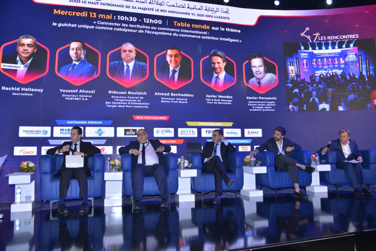 Panel of five men in suits seated on a blue stage during a formal conference, with a large backdrop of portraits and event text behind them.