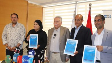 Five adults stand at a press conference, two men in suits and a woman wearing a hijab hold blue posters; microphones on the table and flags behind them.