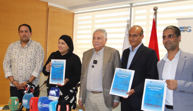 Five adults stand at a press conference, two men in suits and a woman wearing a hijab hold blue posters; microphones on the table and flags behind them.