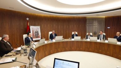 A group of professionals in suits sit around a large curved wooden conference table in a modern meeting room, with laptops and documents in front of them. A Moroccan flag and a framed portrait are visible in the background, suggesting a formal government or corporate meeting.