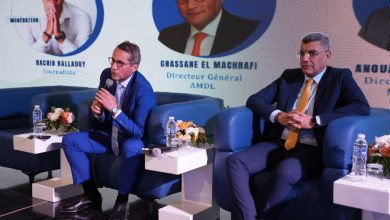Two businessmen in blue suits sit on a panel stage, speaking into a microphone, with floral centerpieces on small tables nearby.