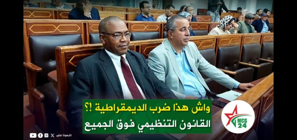 مفتاح يتنقد عقد اجتماعات اللجان بالمقاطعات تزامنا مع توقيت دورات المجلس Two men in business attire sit in a formal assembly hall; an Arabic news banner runs across the bottom and a round logo appears at the bottom-right.
