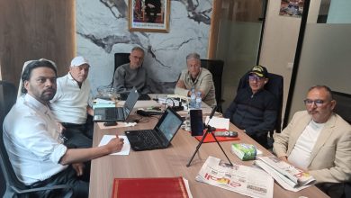 Six men sit around a conference table with laptops and papers in a modern office; a framed portrait hangs on marble wall behind them.