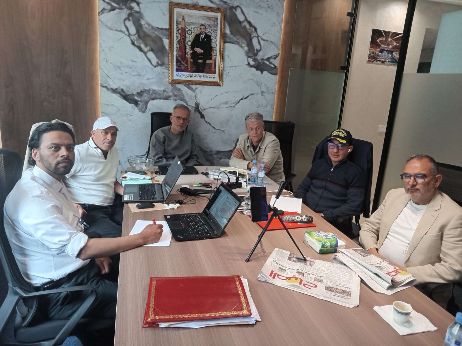 Six men sit around a conference table with laptops and papers in a modern office; a framed portrait hangs on marble wall behind them.