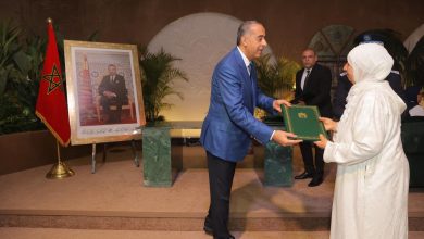 A man in a blue suit presents a green ceremonial folder to a woman in white traditional dress at an awards ceremony, with a Moroccan flag and framed portrait in the background due to formal event setting.