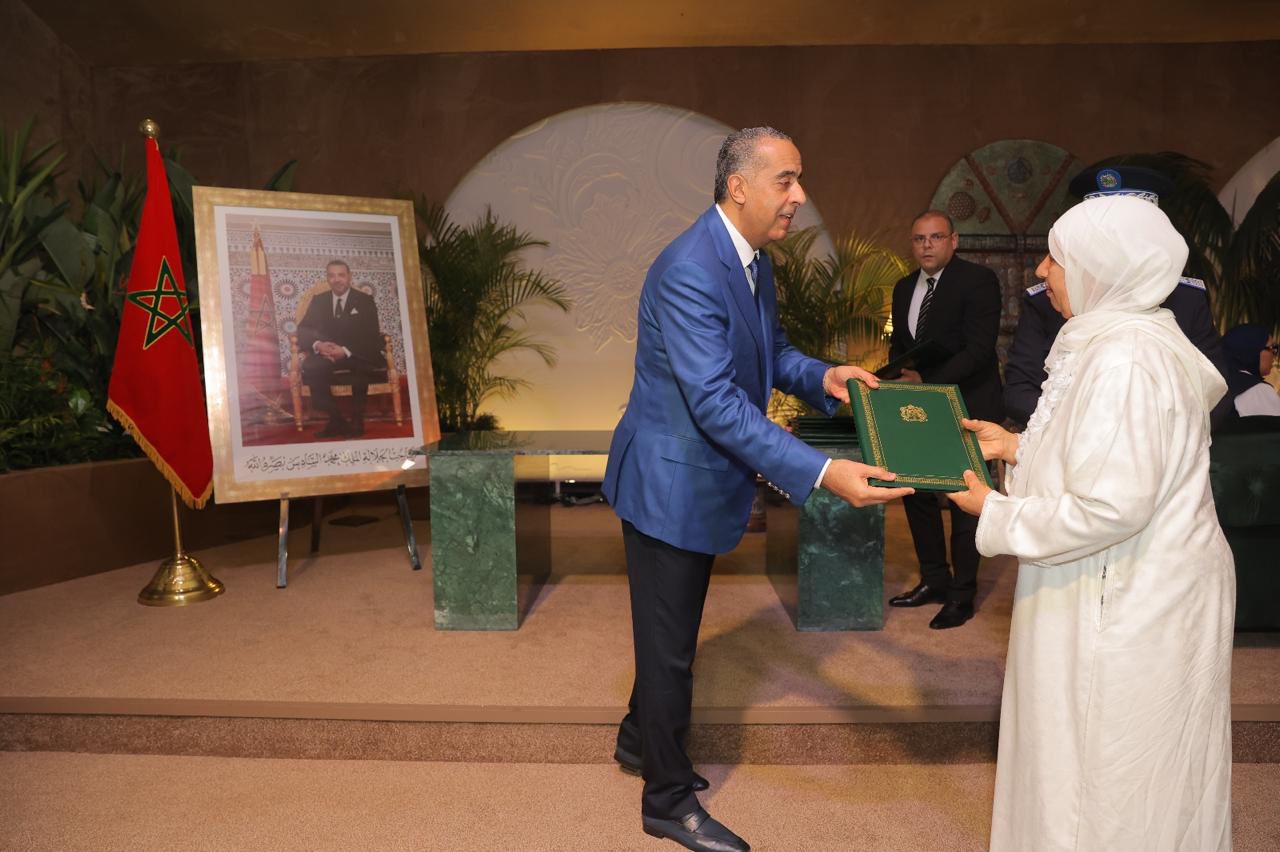 WhatsApp Image 2026-05-19 at 125745 1 | MCG24 | موقع إلكتروني مغربي شامل متجدد على مدار الساعة A man in a blue suit presents a green ceremonial folder to a woman in white traditional dress at an awards ceremony, with a Moroccan flag and framed portrait in the background due to formal event setting.