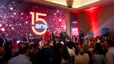 A crowded celebration on stage with speakers in red and black suits, a bright screen showing '15 ans' and confetti-like lights in the background.
