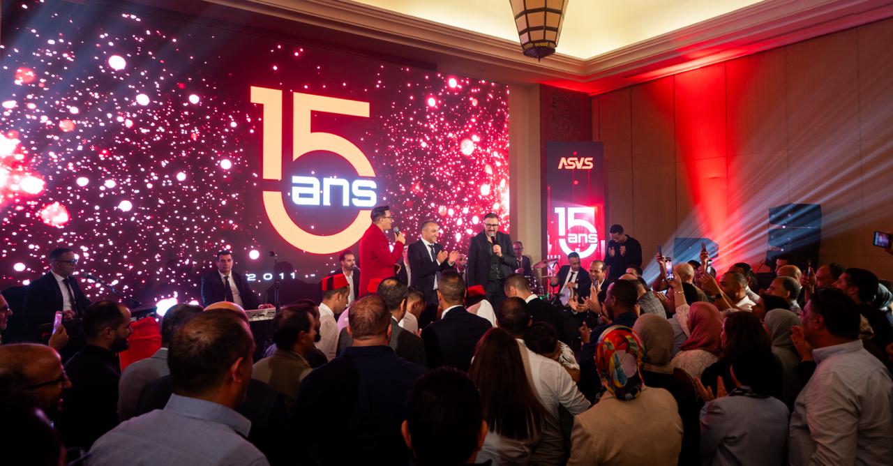 A crowded celebration on stage with speakers in red and black suits, a bright screen showing '15 ans' and confetti-like lights in the background.