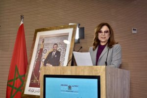 Woman in a gray blazer and purple blouse lecturing at a wooden podium, with a framed portrait and Moroccan flag nearby.