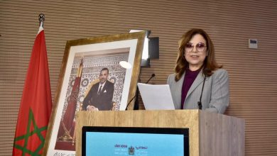 WhatsApp Image 2026-05-19 at 182532 | MCG24 | موقع إلكتروني مغربي شامل متجدد على مدار الساعة Woman in a gray blazer and purple blouse lecturing at a wooden podium, with a framed portrait and Moroccan flag nearby.