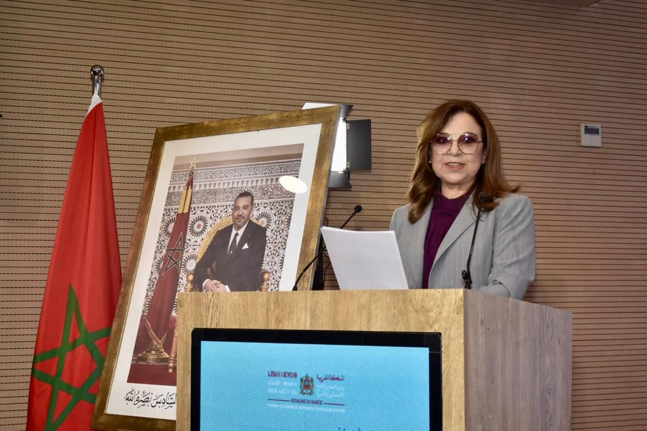 Woman in a gray blazer and purple blouse lecturing at a wooden podium, with a framed portrait and Moroccan flag nearby.