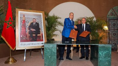 Two men in suits shake hands while holding signed documents during a formal ceremony, with the Moroccan flag and a large framed portrait nearby.