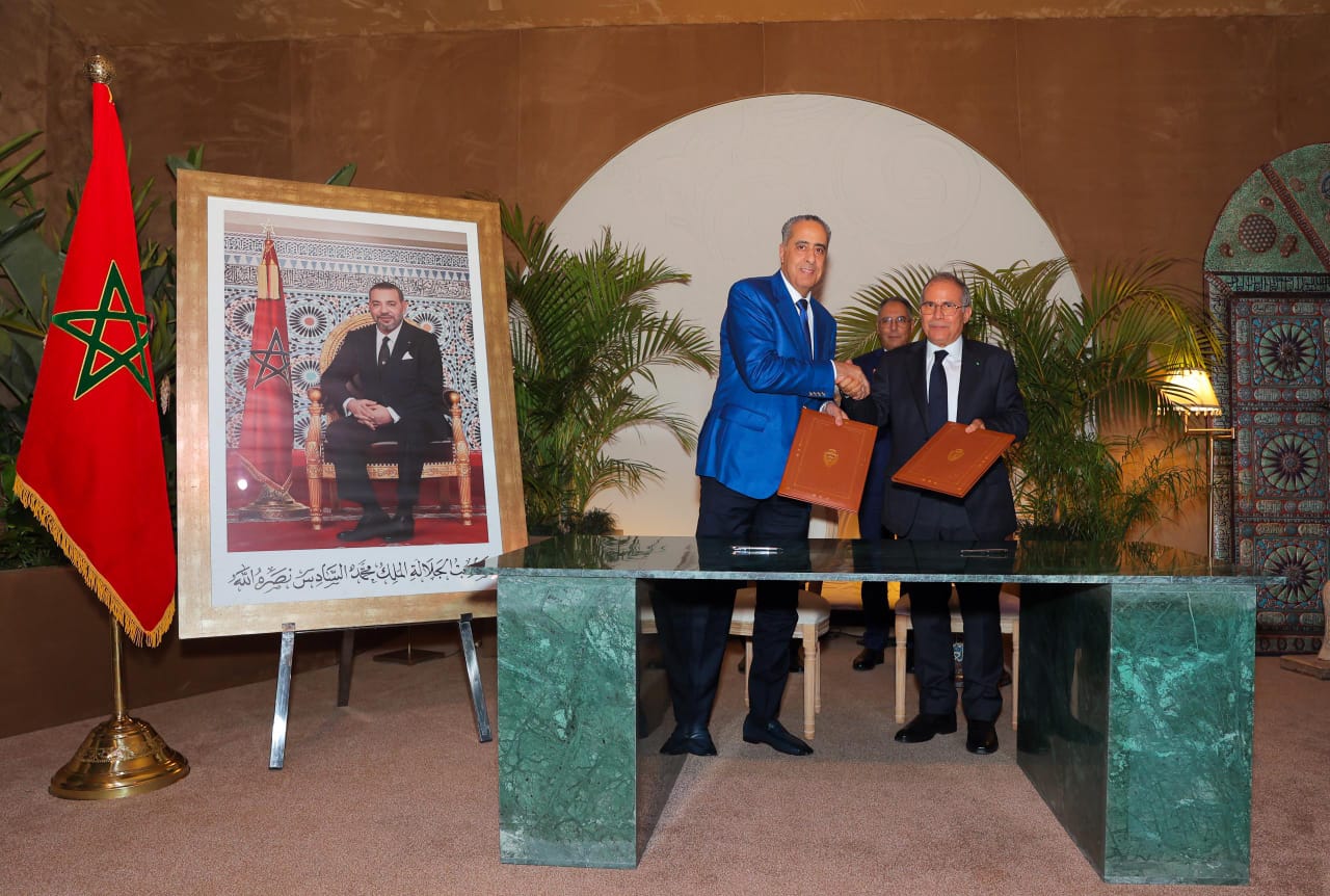 Two men in suits shake hands while holding signed documents during a formal ceremony, with the Moroccan flag and a large framed portrait nearby.