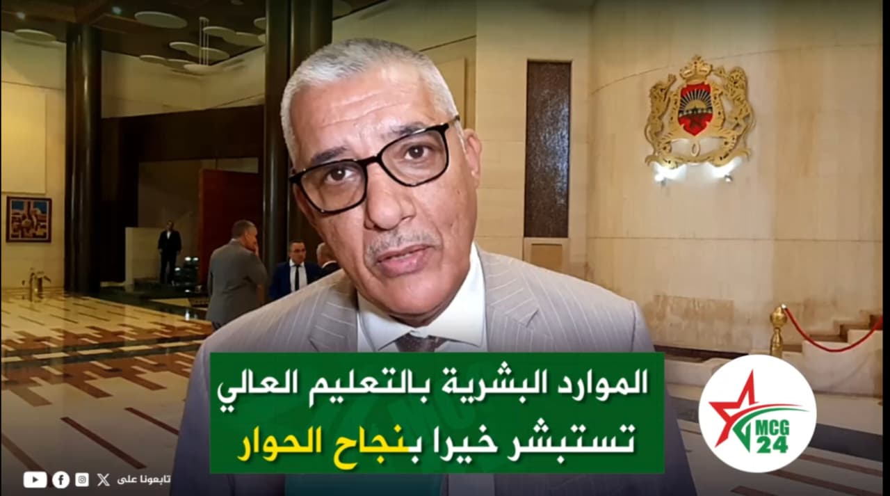 Man in a light gray suit and black glasses speaks in a lobby with a green Arabic caption overlay about higher education resources and dialogue, and a logo reading MC G24.