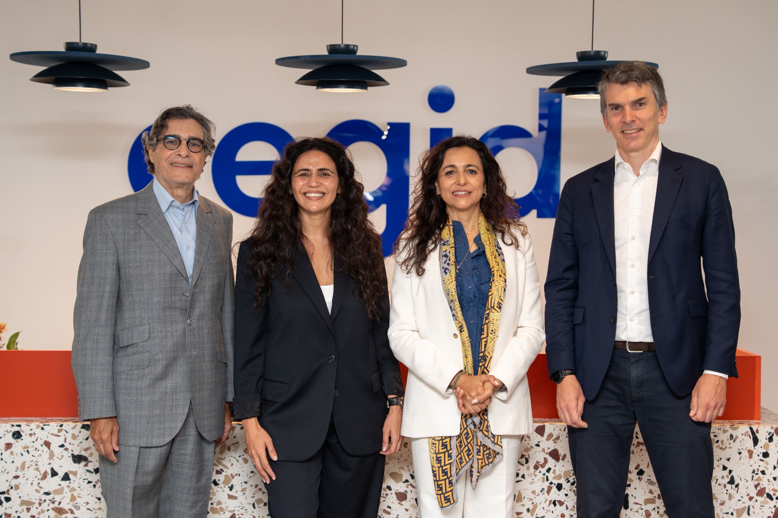 Four professionals in business attire smile for a group photo indoors in front of a blue logo.