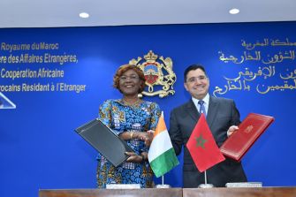 Two diplomats stand at a signing ceremony with a blue backdrop, each holding a document; Moroccan flag and another national flag are displayed on the table behind them.