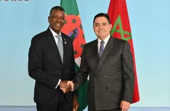 Two men in dark suits shake hands in front of a blue backdrop with flags behind them, signaling a formal meeting or agreement.