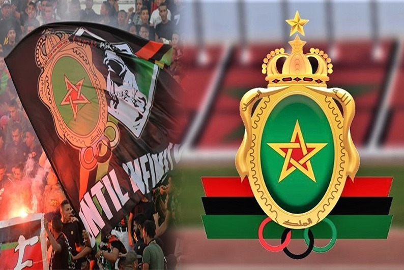 Crowd of Moroccan football fans waving a flag beside the Royal Moroccan Football Federation crest in a stadium background.