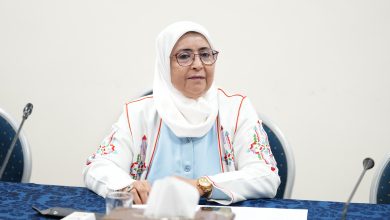 Woman wearing a white hijab and glasses sits at a table with papers, a microphone, and chairs in a meeting room, looking forward slightly to her left.