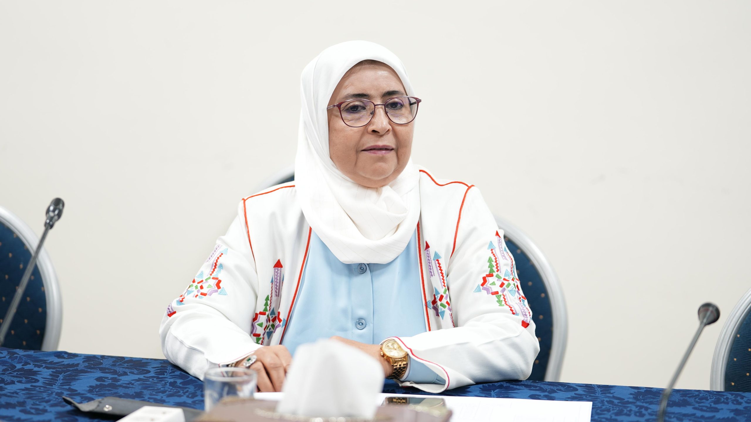 Woman wearing a white hijab and glasses sits at a table with papers, a microphone, and chairs in a meeting room, looking forward slightly to her left.