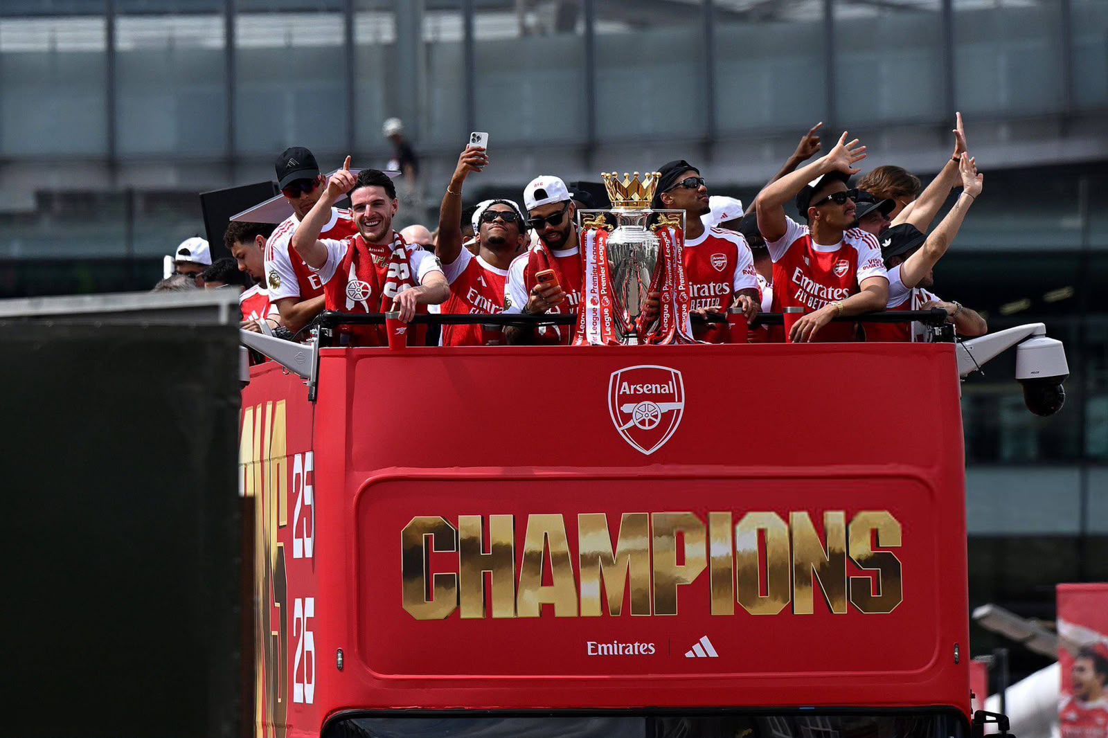 Arsenal players celebrate on a red double-decker bus, raising their arms around the Premier League trophy.