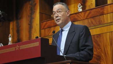 A man in a dark suit and blue tie speaks at a wooden podium with microphones in front of a wood-paneled backdrop.