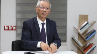 Older man in a dark pinstripe suit and purple tie sits at a white table, pointing at documents during a meeting.