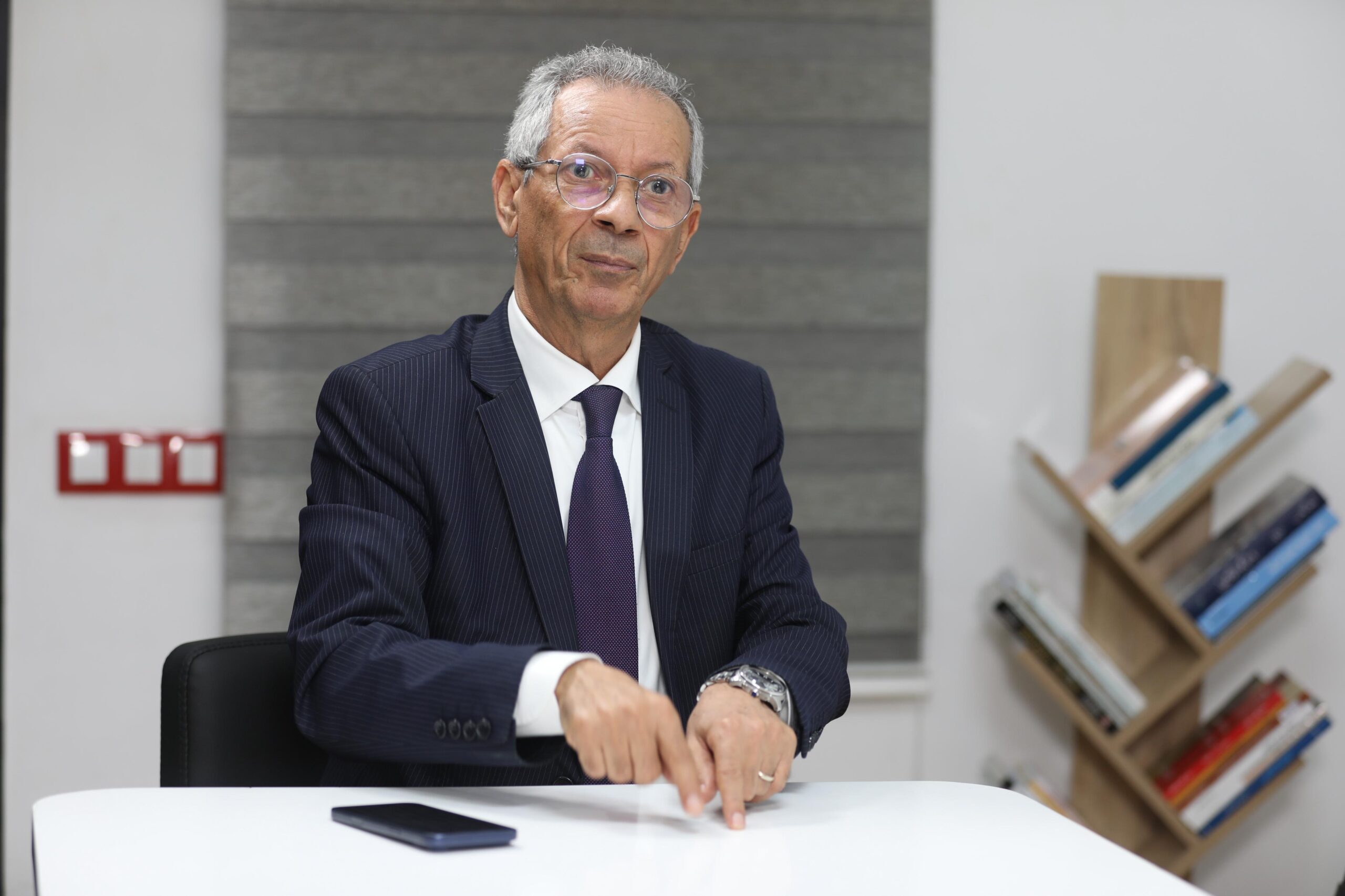 Older man in a dark pinstripe suit and purple tie sits at a white table, pointing at documents during a meeting.