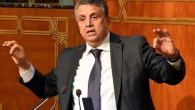 Man in a dark pinstripe suit giving a speech at a podium with two microphones, gesturing with his hands.
