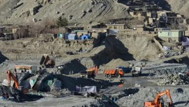 Open-pit quarry with orange excavators, dump trucks, and temporary shelters scattered on a rocky hillside.