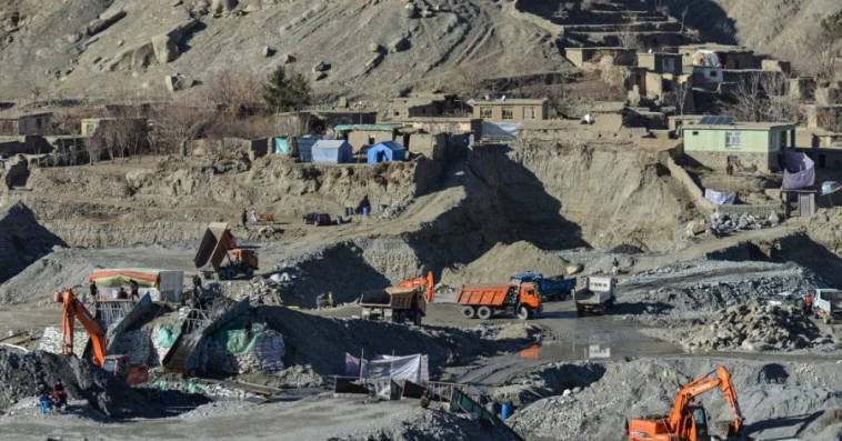 Open-pit quarry with orange excavators, dump trucks, and temporary shelters scattered on a rocky hillside.