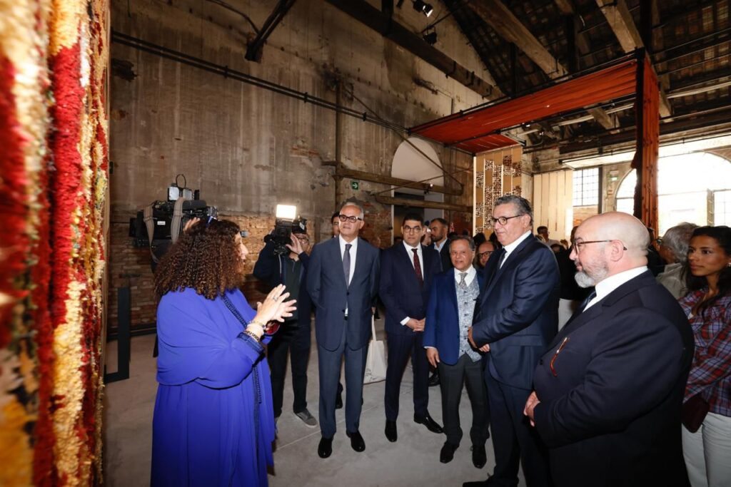 Woman in a blue dress speaks to a group of suited professionals in an industrial textile gallery, with a colorful rug on the left.