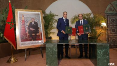 akhbar | MCG24 | موقع إلكتروني مغربي شامل متجدد على مدار الساعة Two men in suits exchange signed documents at a formal ceremony, with a Moroccan flag and a large framed portrait nearby.