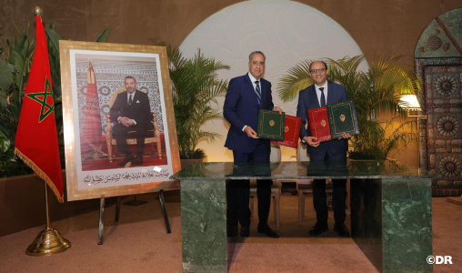 Two men in suits exchange signed documents at a formal ceremony, with a Moroccan flag and a large framed portrait nearby.