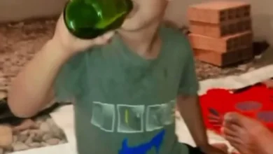 alcool-mineur | MCG24 | موقع إلكتروني مغربي شامل متجدد على مدار الساعة Person in a gray shirt drinks from a large green bottle indoors, with a stack of red bricks in the background.