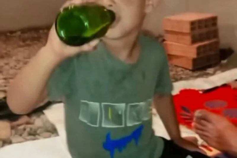 alcool-mineur | MCG24 | موقع إلكتروني مغربي شامل متجدد على مدار الساعة Person in a gray shirt drinks from a large green bottle indoors, with a stack of red bricks in the background.